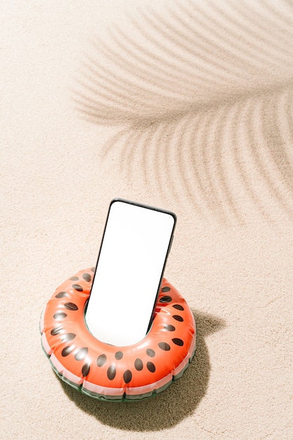 Foto sandía inflable con forma de teléfono móvil en el interior sobre la arena de la playa tropical con sombras de hojas de cocotero en verano