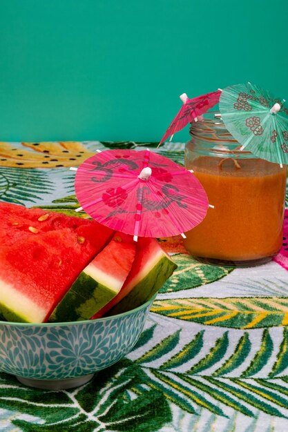 Sandía recién cortada con un pequeño paraguas, un vaso de gazpacho en una mesa con palmeras.