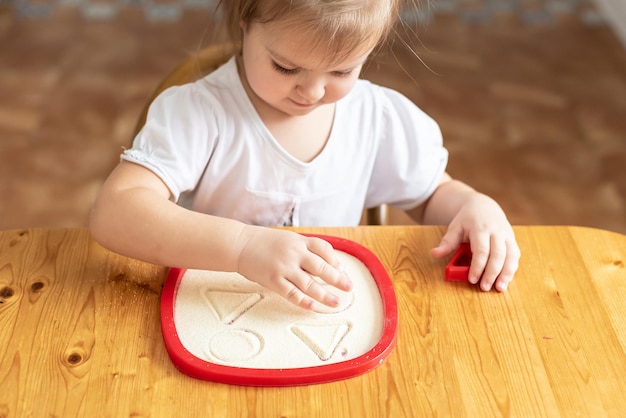 Sandkunsttherapie kleines Mädchen auf einem Tisch mit Sand gemalt Sinnesspiele zu Hause Spielen im Grieß in der Küche Entwicklung der Feinmotorik Aktivitäten Montessori