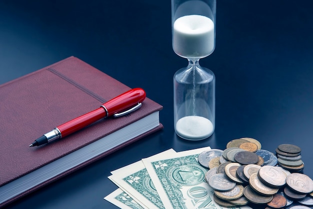 Sanduhr, Geld, Stift und Notizbuch liegen auf dem Tisch