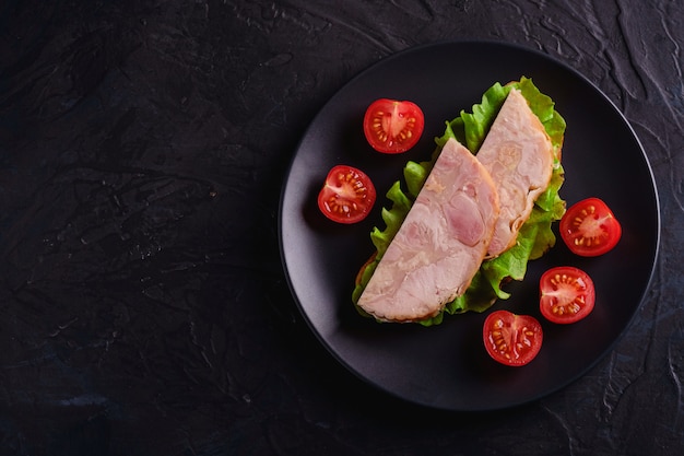 Sanduíche com carne de presunto de peru, salada verde e fatias de tomate cereja fresco em chapa preta, mesa texturizada escura, vista superior