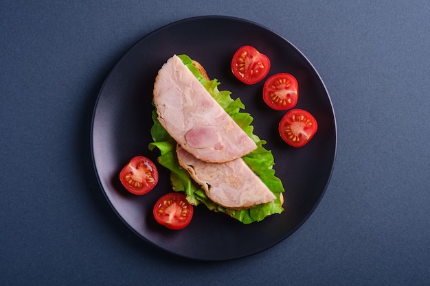 Sanduíche com carne de presunto de peru, salada verde e fatias de tomate cereja fresco na mesa preta, azul mesa mínima, vista superior