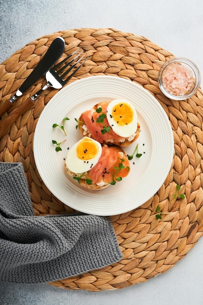 Sanduíche com coberturas deliciosas em cima de ovos de salmão defumado ervas e microgreens rabanete sementes de gergelim preto sobre prato branco sobre fundo de concreto Superalimento saudável sanduíche aberto Vista superior