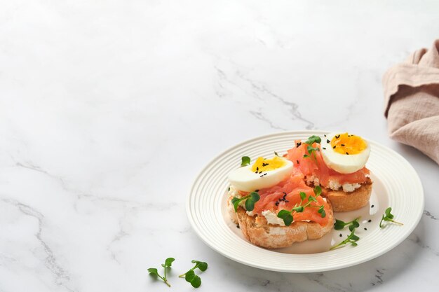 Sanduíche com coberturas deliciosas ovos de salmão defumado ervas e microgreens rabanete sementes de gergelim preto sobre prato branco no fundo da mesa de mármore branco Superalimento saudável sanduíche aberto Vista superior