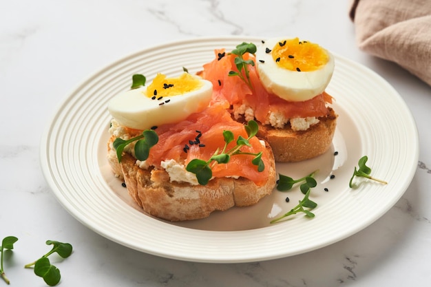 Sanduíche com coberturas deliciosas ovos de salmão defumado ervas e microgreens rabanete sementes de gergelim preto sobre prato branco no fundo da mesa de mármore branco Superalimento saudável sanduíche aberto Vista superior