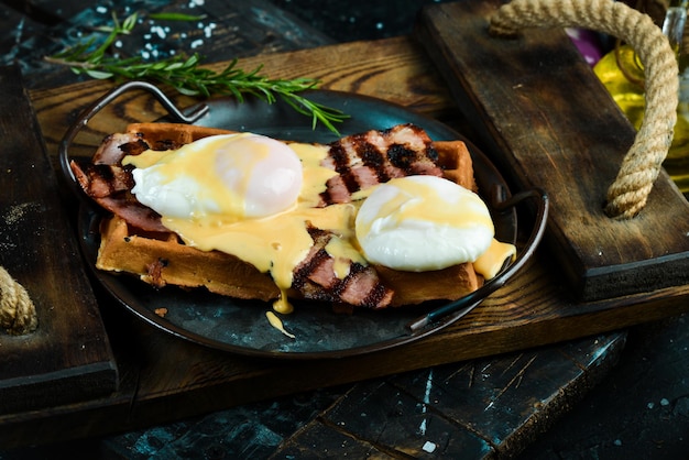 Sanduíche com ovo escalfado e bacon em um prato nas mãos de um chef em um fundo preto. waffles belgas.