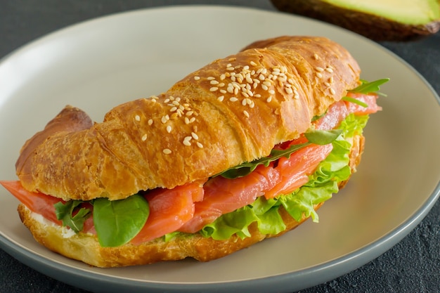 Sanduíche de croissant com salmão, ricota e rúcula em um prato em um fundo escuro.