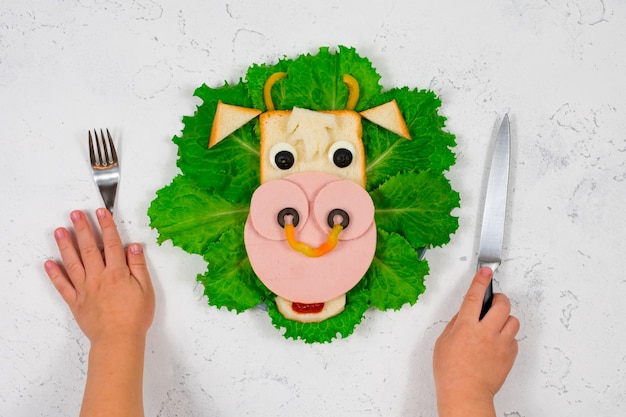 Foto sanduíche engraçado com o símbolo do touro comestível de 2021 feito de torradas e linguiça em folhas verdes de alface. as mãos das crianças estão segurando um garfo e uma faca. ideia de pequeno-almoço para crianças.
