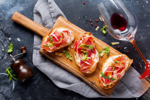 Sanduíches abertos de presunto, rúcula e queijo duro, servidos em uma estante de madeira com um copo de vinho tinto em uma superfície de concreto velha e escura