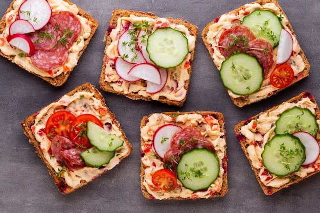 Sanduíches com cream cheese, legumes e salame. Sanduíches com pepino, rabanete, tomate, salame em um fundo cinza, vista superior. Postura plana.