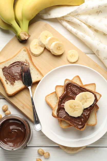Sanduíches com manteiga de chocolate, bananas e avelãs sobre uma mesa de madeira branca.