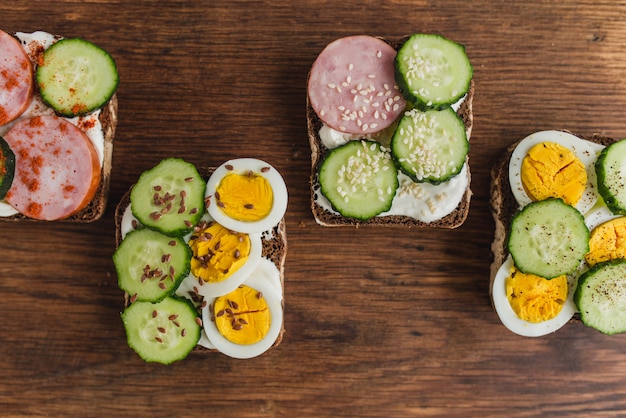 Foto sanduíches com pão de centeio, requeijão, pepino, ovo, presunto, especiarias e sementes. conjunto de deliciosos sanduíches em uma prancha velha rústica. vista superior, estilo liso leigo.