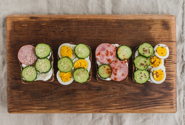 Foto sanduíches com pão de centeio, requeijão, pepino, ovo, presunto, especiarias e sementes. conjunto de deliciosos sanduíches em uma prancha velha rústica. vista superior, estilo liso leigo.