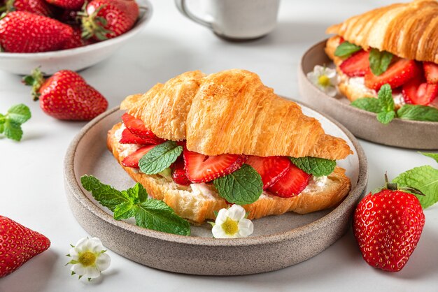 Sanduíches de croissant com morangos maduros frescos e cream cheese em um prato com uma xícara de café no saboroso café da manhã na mesa branca