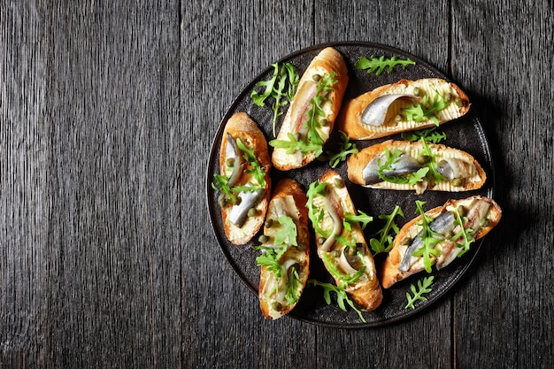 Sanduíches de manteiga de anchova com folhas frescas de salada de rúcula, alcaparras em uma travessa preta em uma mesa de madeira rústica, vista horizontal de cima, postura plana, espaço livre