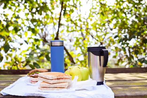 Sanduíches e duas garrafas térmicas com chá e café em uma mesa de madeira no parque em um fundo de folhas verdes, uma árvore e luz solar. Conceito de turismo e viagens. Copie o espaço