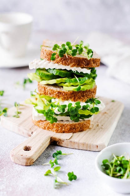 Sándwich de aguacate, pepino y queso feta decorado con microgreens y pan multigrano en un sencillo soporte de madera para un desayuno saludable. Enfoque selectivo