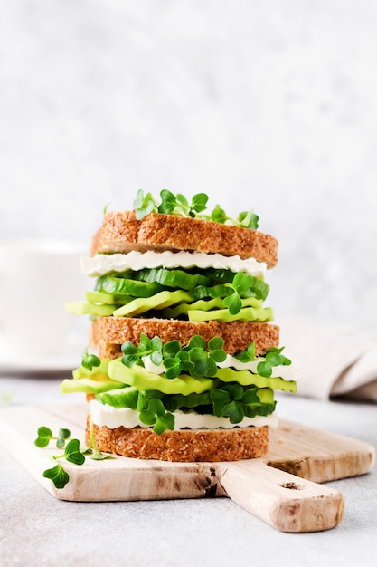 Sándwich de aguacate, pepino y queso feta decorado con microverduras y pan multigrano en un sencillo soporte de madera para un desayuno saludable