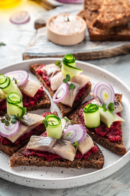 Sándwich con arenque, remolacha, pepino verde, cebolla y microverde, tostadas de pan oscuro, tradición danesa, cocina escandinava, imagen vertical, vista superior