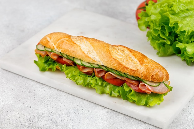 Sándwich de baguette fresco con jamón tomate lechuga pepino sobre una placa de mármol y fondo gris