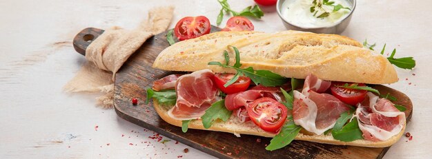 Sándwich de baguette con jamón de jamón, tomates de cereza y rúcula en un tablero Con fondo blanco vista panorámica desde arriba