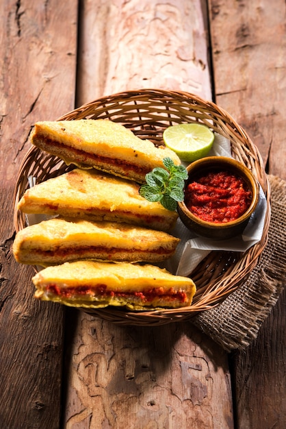 Sandwich Bread Pakora oder Pakoda (Dreiecksform) serviert mit Tomatenketchup, Chutney, grünen Chili- und Zwiebelscheiben, beliebter indischer Snack zur Teezeit. Selektiver Fokus