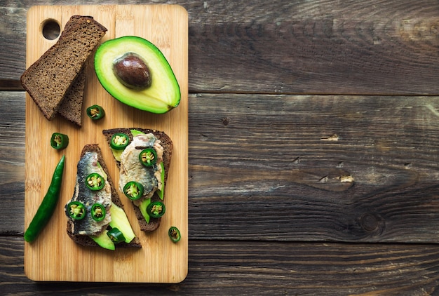 Foto sándwich de cara abierta con aguacate, sardina y jalapeño sobre pan de centeno oscuro sobre madera rústica. refrigerio saludable.