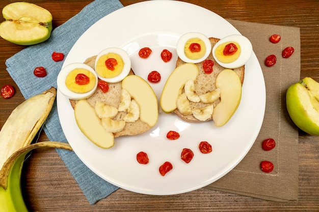 Sándwich de cara de búho divertido pan tostado con huevos de pollo manzana plátano frutos de bayas secas en plato Niños lindos niños postre dulce desayuno almuerzo comida arte sobre fondo de madera vista superior