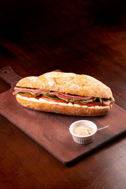 Foto sándwich de carpaccio ahumado con tomate de rúcula y queso gruiere en baguette italiana sobre tabla de madera y vista superior de fondo oscuro con retrato de mayonesa