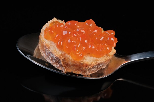 Un sándwich con caviar de salmón chum rojo salado se encuentra en una cuchara de metal sobre negro