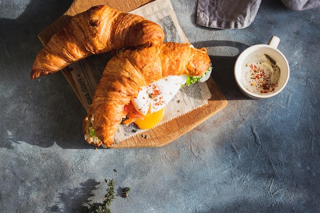 Sándwich de croissant con huevo escalfado, hojas de ensalada, salmón y requesón cortado en tabla de cortar con café. concepto de desayuno