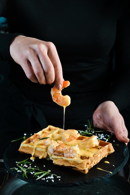 Sándwich de gofres belgas con camarones y salsa de queso en un plato en manos de un chef Sobre un fondo negro