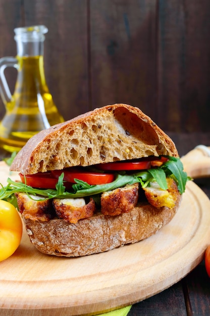 Sándwich grande con trozos de carne, rúcula, tomate, ciabatta de cereales en la tabla de cortar sobre la mesa de madera oscura.