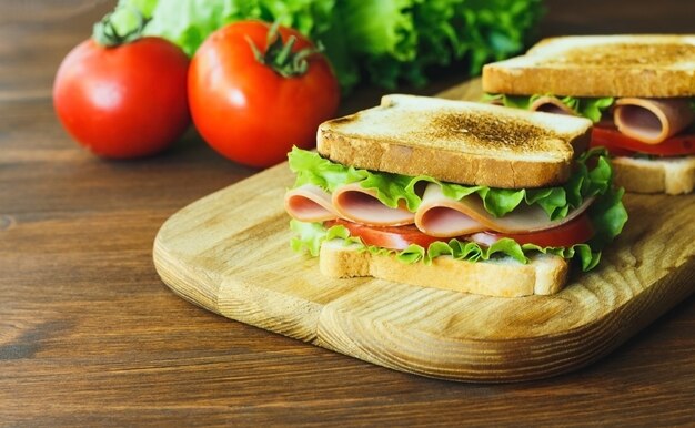 Sándwich de jamón, lechuga verde y tomate sobre una placa de cocina de madera marrón