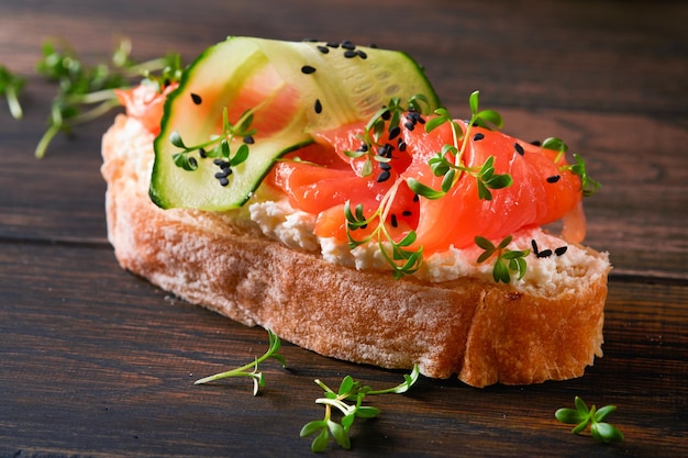 Sandwich Lachstoast mit Frischkäse, Gurke, schwarzem Sesam und Microgreens auf altem Holztischhintergrund Meeresfrüchte Gesunde Lebensmittel Fotografie in zurückhaltender Ansicht von oben