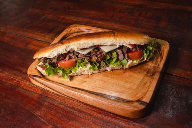 Sándwich largo con carne, tomate y lechuga en una tabla sobre una mesa de madera.