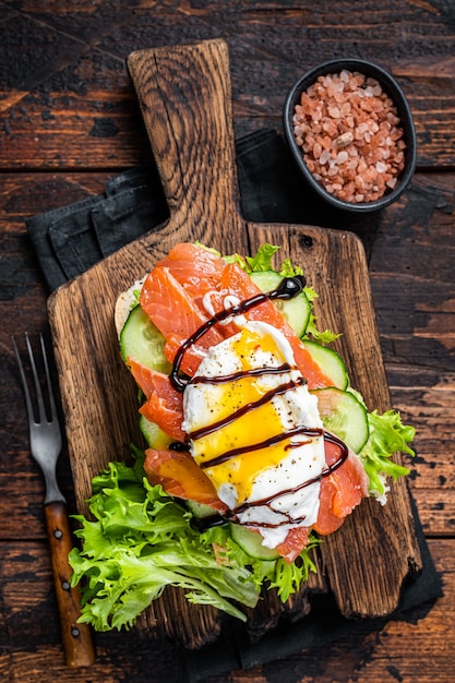 Sandwich mit geräuchertem Lachs mit Benedict-Ei und Avocado auf Brot. Dunkler hölzerner Hintergrund. Ansicht von oben.