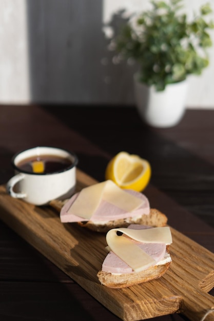 Sandwich mit geschnittener Schinkenbutter und Käse auf Holzschneidebrett in der Nähe einer Tasse Tee mit Zitrone