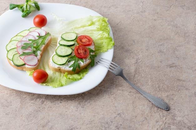 Sandwich mit Hüttenkäse, Gurken und Kirschtomaten und Rettich auf einem weißen Teller auf dem Tisch. Teller mit einer Gabel auf dem Tisch mit einem weißen Teller mit Gemüsesalat.