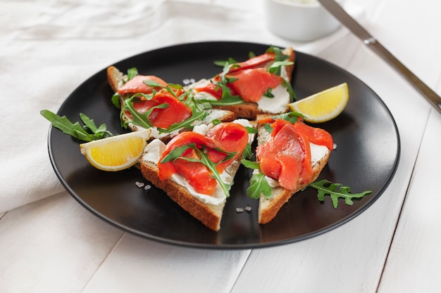 Sandwich mit Lachs, Käse, Rucola und Zitrone auf einem schwarzen Teller auf einem weißen Tisch