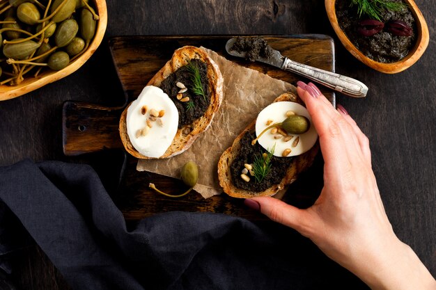 Sandwich mit Scheibe Mozzarella-Käse und Tapenade, Kapern auf dunklem rustikalem Tischhintergrund. Weibliche Hand, die Sandwich hält. Traditionelles Gericht der Provence. Ansicht von oben