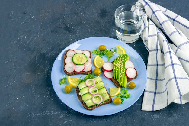 Sandwich mit verschiedenen Belägen Avocado, Gurke, Rettich auf Teller mit einem Glas Wasser auf dunklem Hintergrund