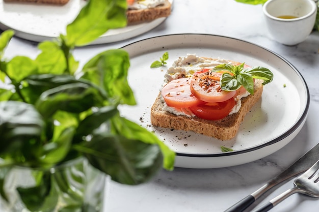 Foto sándwich o tostada con tomates, queso crema, aceite de oliva y albahaca en un plato con fondo de mármol blanco