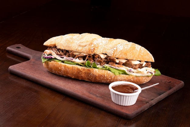 Sándwich de pechuga de ternera de comida rápida con rúcula y ensalada de col en pan baguette sobre tabla de madera con salsa barbacoa fondo de ángulo oscuro