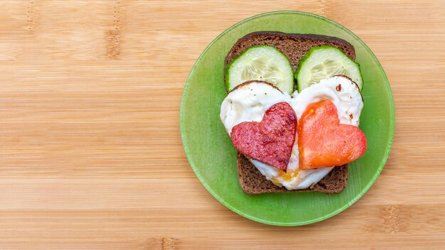 Sándwich de pepino, huevos revueltos en forma de corazón con salchichas y tomate en forma de corazón en una placa verde