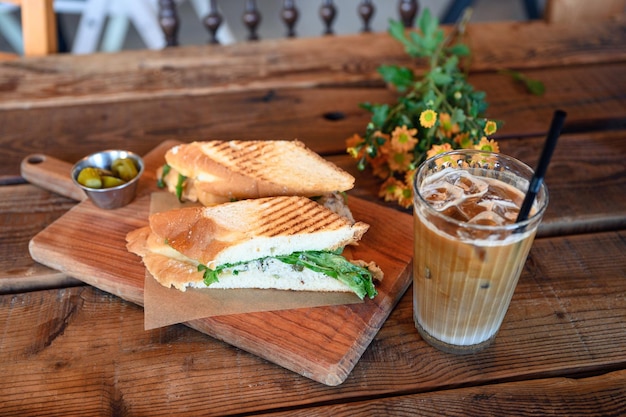 Sándwich de pollo con café helado y decoración floral en mesa de madera