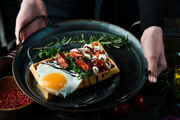 Sándwich con queso de huevo, pescado salado y rúcula en un plato en manos de un chef sobre un fondo rústico de madera Gofres belgas Sobre un fondo negro