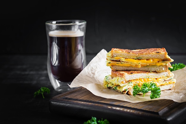 Sándwich de queso tostado y huevos revueltos sobre papel con taza de café sobre tabla de madera. Fondo oscuro.