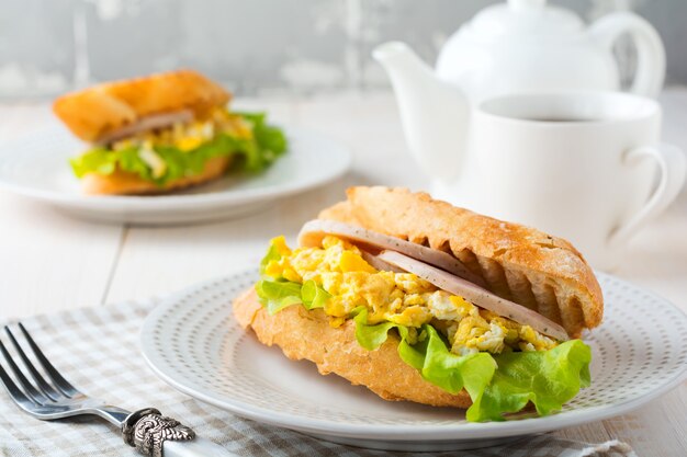 Sandwich relleno de huevos revueltos, jamón y hojas de lechuga sobre un fondo de madera clara. Enfoque selectivo.