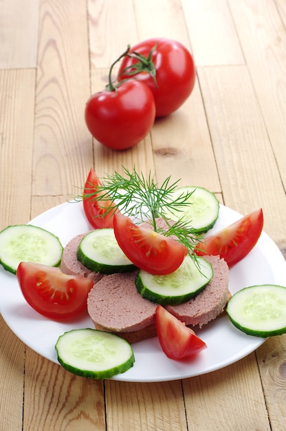 Sándwich con salchicha de hígado, pepino fresco y tomate en un plato sobre una mesa de madera